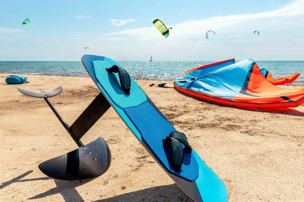 Close-up von Tragflächenboot, Surfbrett und Kite-Ausrüstung am Sandstrand. Wassersport vor Ort an einem hellen, sonnigen Tag am Meer, mit vielen Kitern, die reiten. Surfschule und Camp. Aktive Reise und Sportkonzept.