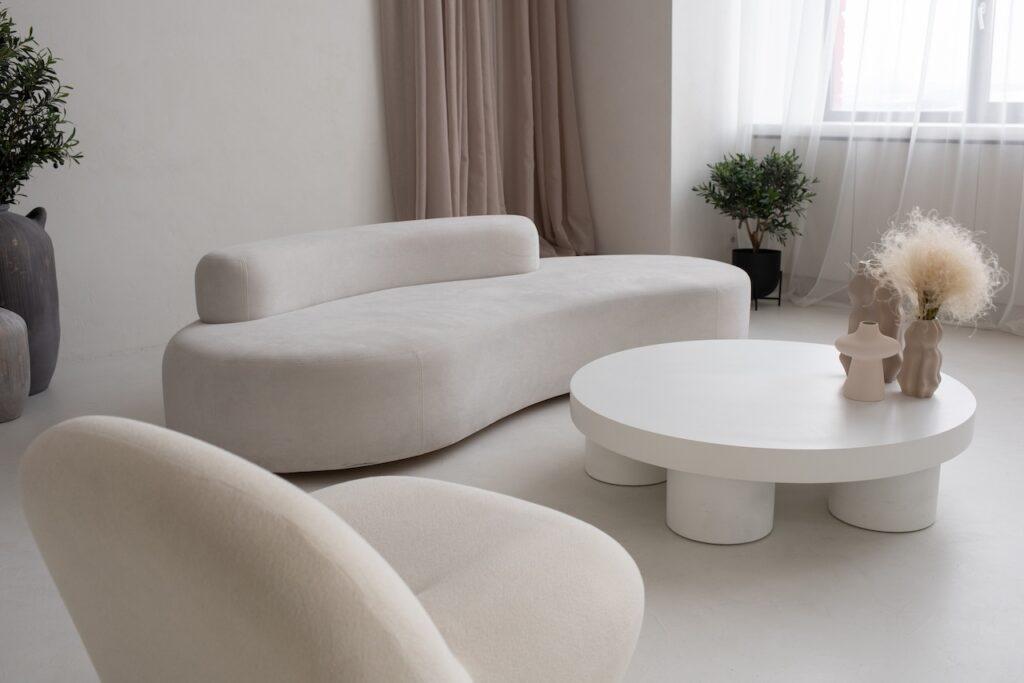 Heller, minimalistischer Couchtisch mit Deko aus Pampasgras und einer Zeitschrift in einem Wohnzimmer mit grauem Sofa und grünen Kissen, dekoriert mit Spiegel und Regalen im Hintergrund.