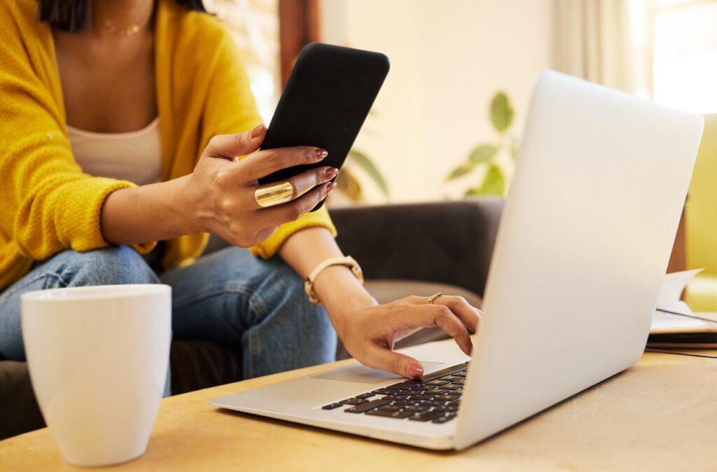 Nahaufnahme einer Frau, die am Couchtisch sitzt, mit einem Smartphone in der einen und einem Laptop in der anderen Hand, während sie Reisepläne erstellt.