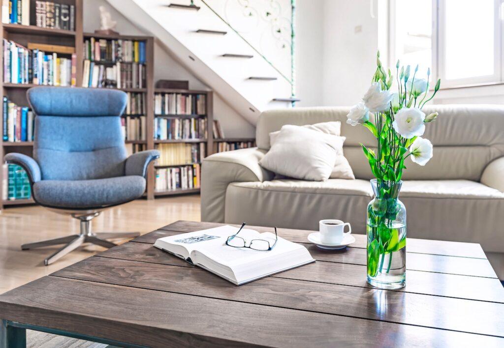 Wohnzimmer mit einem Couchtisch aus dunklem Holz, darauf eine offene Brille, ein Buch, eine Tasse Kaffee und eine Vase mit weißen Blumen, umgeben von einer hellen Ledercouch und einem Bücherregal.