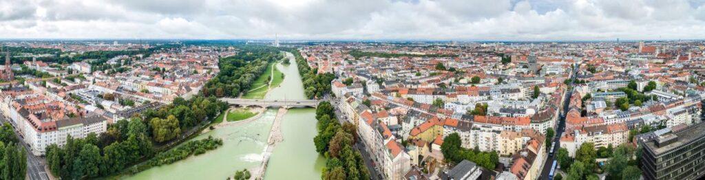 Klassenfahrt München: Panoramaaufnahme mit Isar, Brücken, Bäumen und Stadtgebiet bei bewölktem Himmel